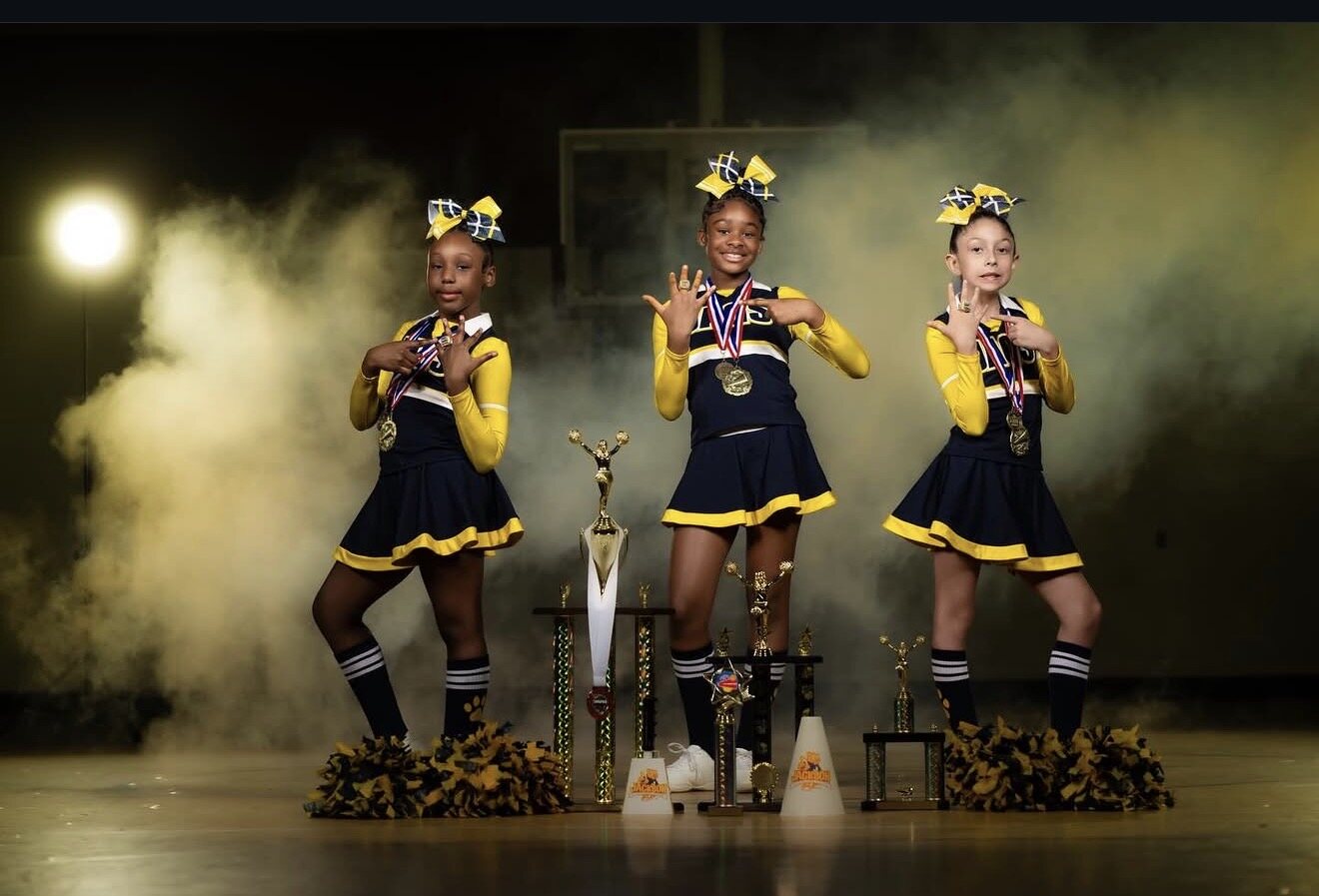 Three cheer athletes in navy and gold uniforms posing with medals and trophies