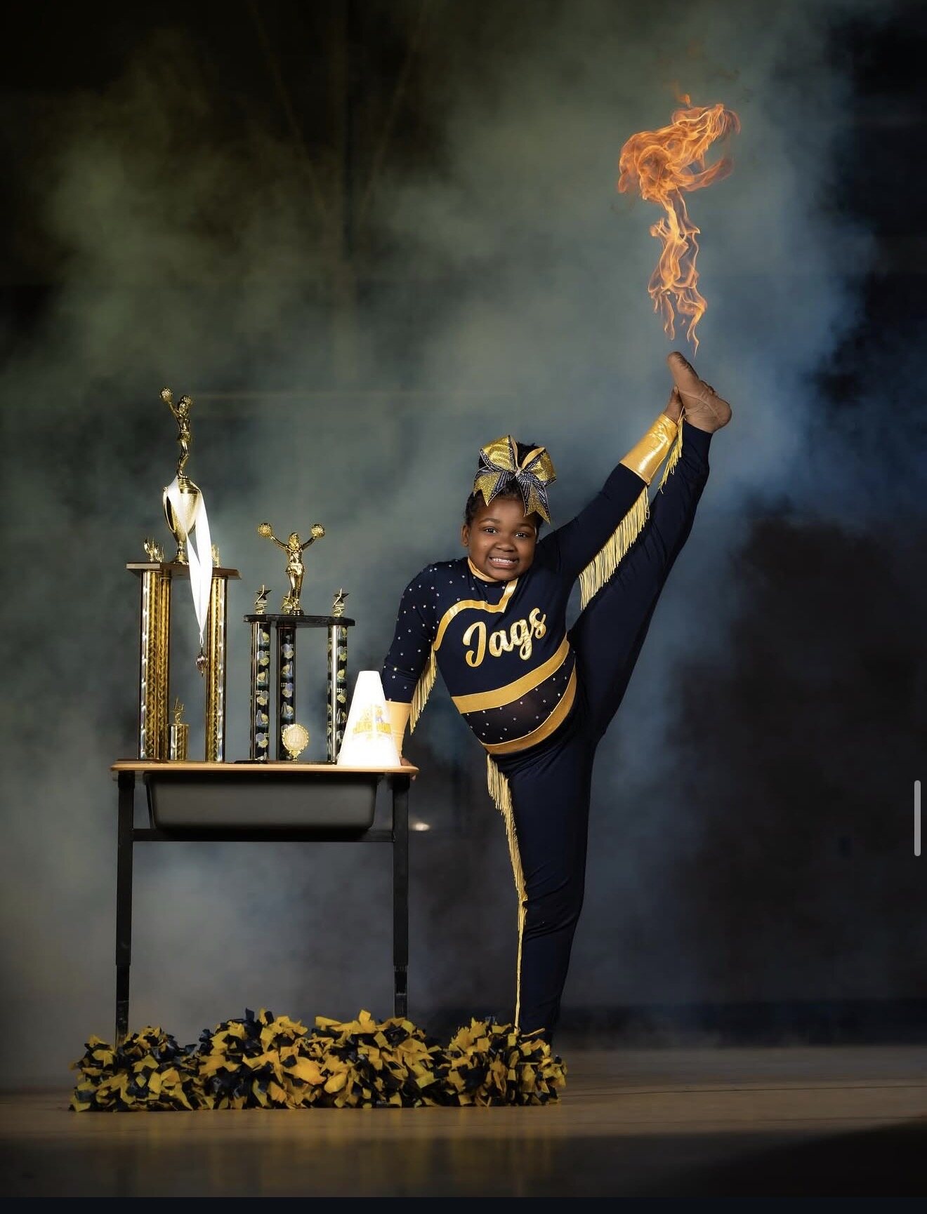 Cheer athlete in a navy and gold Jags uniform performing a heel stretch beside trophies